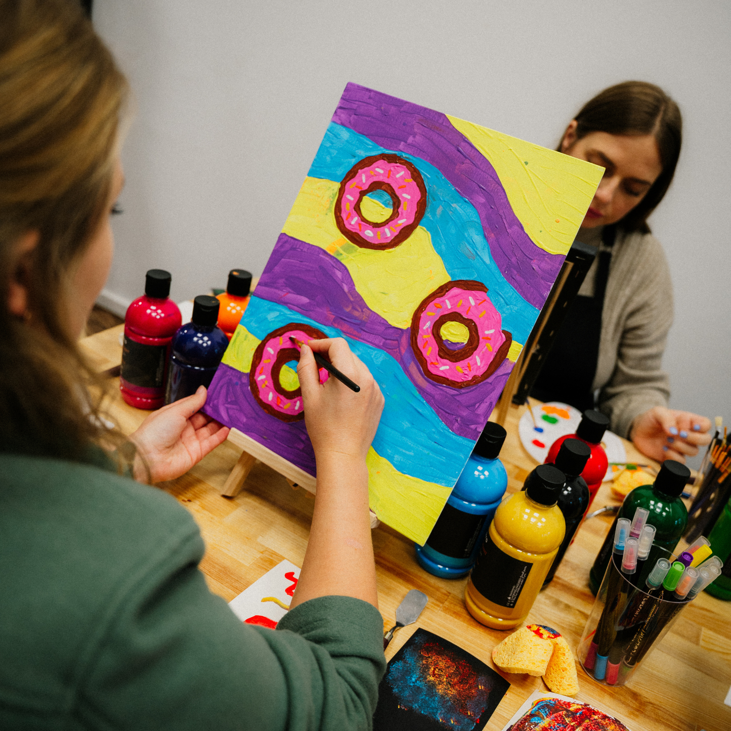 Eine Teilnehmerin des Workshops Malen mit Acrylfarben malt ein buntes Bild mit Wellen und Donuts. Im Hintergrund eine andere Teilnehmerin.