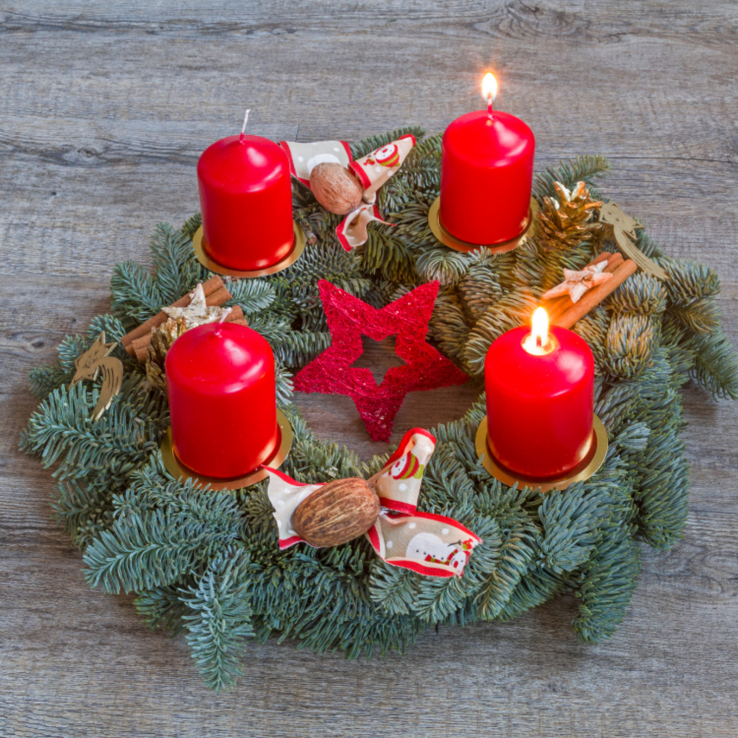 Verzierter Adventskranz mit roten Kerzen auf einem Tisch bei dem zwei Kerzen brennen.