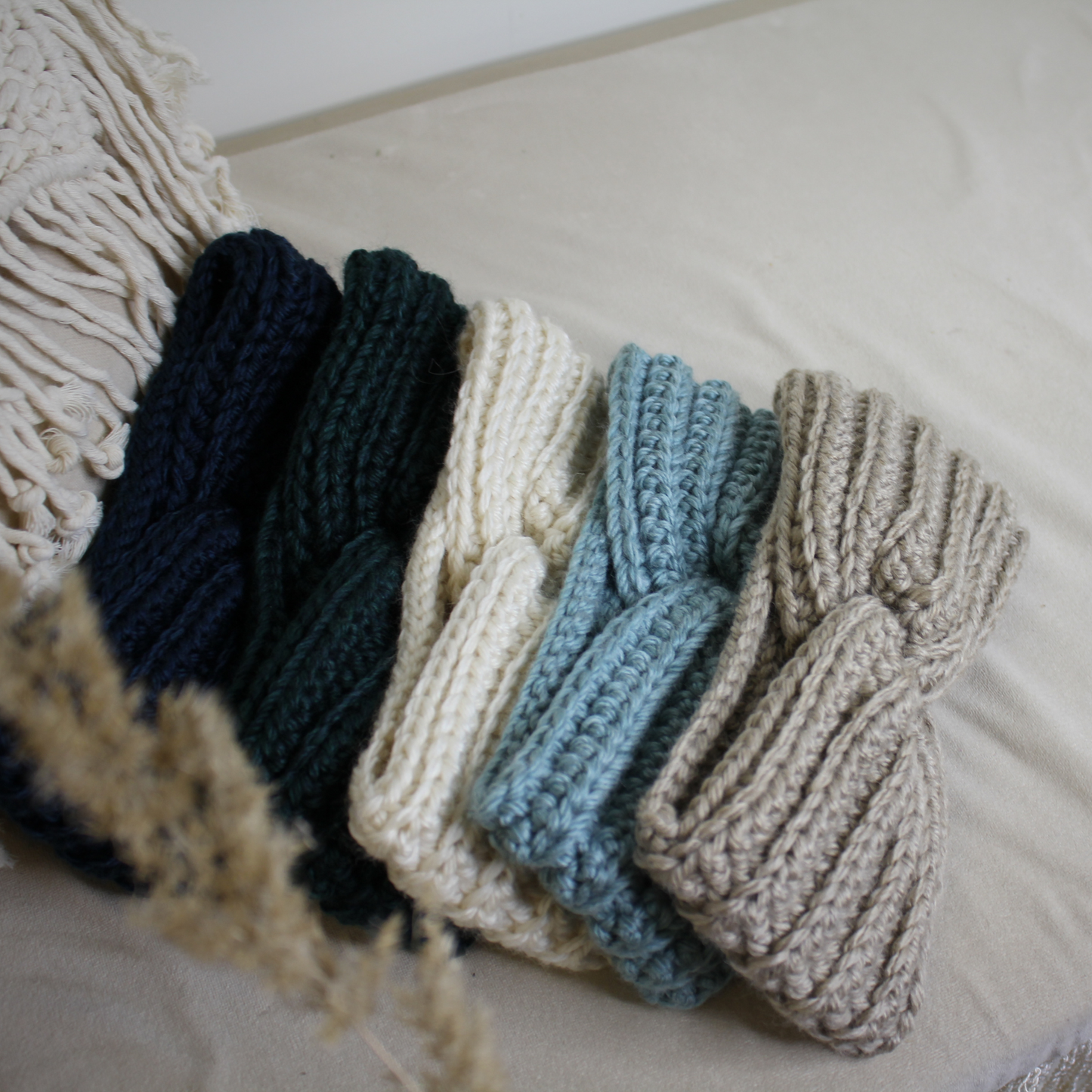 Fünf gestrickte Schals liegen auf einem Tuch mit einer Trockenblume im Hintergrund.