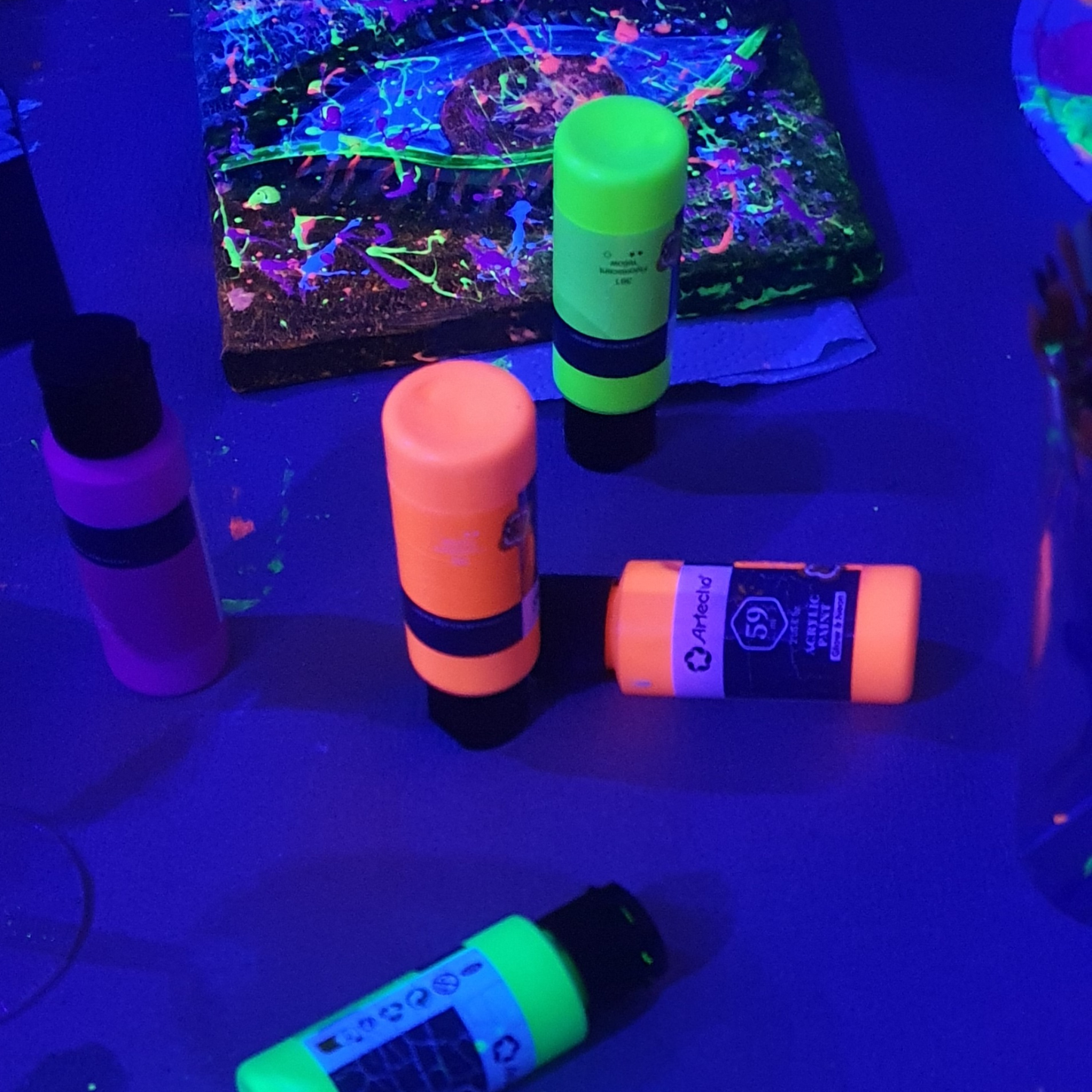 Leuchtende UV-Farbtuben und ein Gemälde mit Neonfarben im Hintergrund bei einem Workshop im Kreativstudio München. Technomusik.