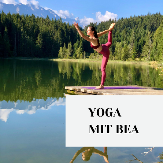 Eine junge Frau steht in Yogaposition auf einem Steganleger an einem See, im Hintergrund die Berge und Wald.