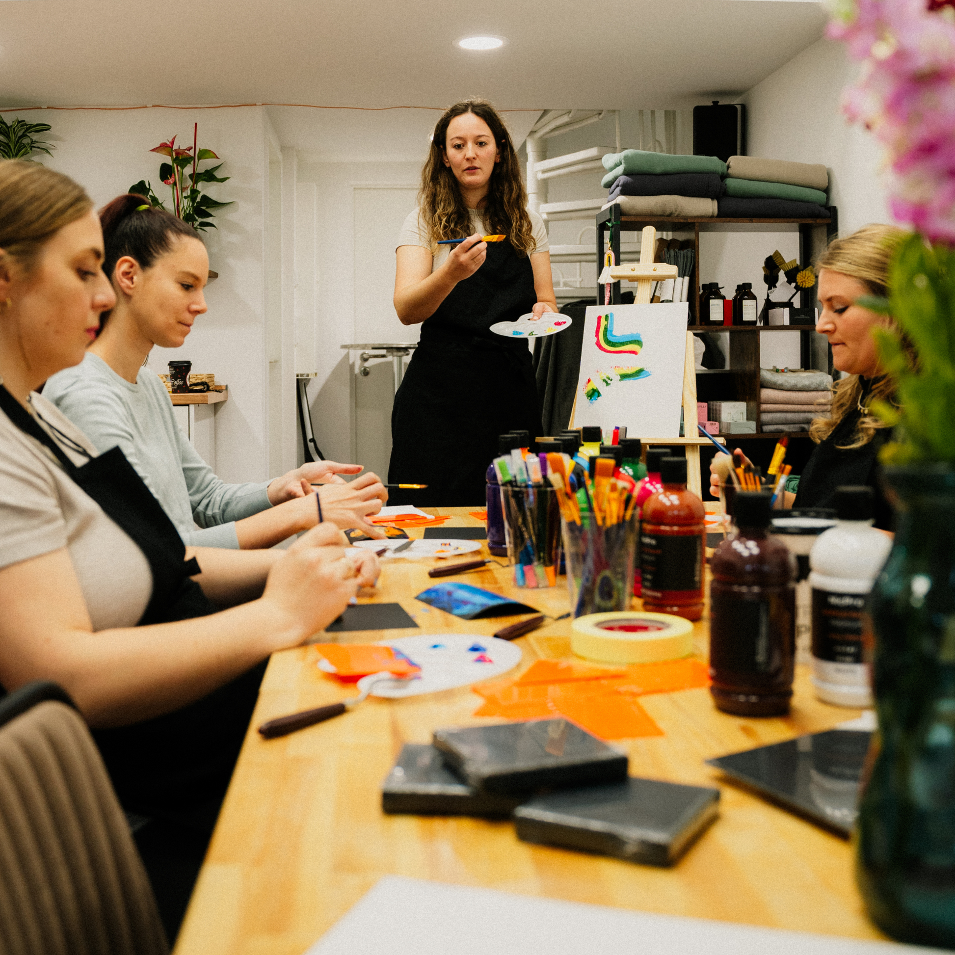Die Kursleiterin leitet die Teilnehmer in einem Acrylfarbenworkshop an. Auf dem Tisch befinden sich zahlreiche Acrylfarben.