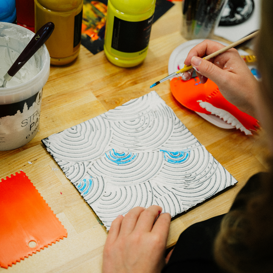 Person hat einen Pinsel in der Hand und erstellt ein Strukturbild im Workshop mit Strukturpaste und Farbe.