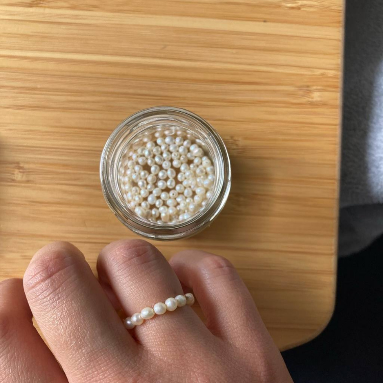 Kleines Glas mit kleinen Süßwasserperlen für einen Workshop und Hand mit einem Fingerring aus weißen Perlen.